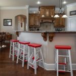 A kitchen with wooden cabinets and granite countertops features four white and red bar stools at a counter. There's a dining area with a wooden table and chairs in the background, and wood flooring throughout.