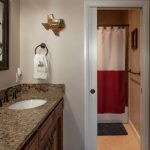 A bathroom features a granite countertop with a sink, a large mirror, and Texas-themed decor, including a Texas-shaped wall hanging and a flag. The adjoining room has a shower with a Texas flag shower curtain.