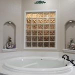 A white oval bathtub is set against a wall with a decorative glass block window. Niches on each side hold seashells and small sculptures. A bronze faucet is on the tub's edge; a white door and window are visible.