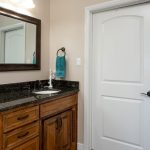 A bathroom with a wooden vanity, black granite countertop, rectangular mirror, towel ring with a teal towel, white door, and beige walls. A light fixture is above the mirror. Part of a bathtub is visible.
