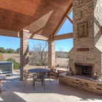 A covered outdoor patio with a stone fireplace, metal table and chairs, wooden picnic table, and a hot tub, overlooking a scenic landscape with trees and open fields under a clear blue sky.