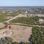 Aerial view of a rural landscape with scattered trees, a round building marked by a red location pin, nearby roads, and distant water bodies under a clear sky.