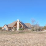 A large stone and brick house with a tall peaked roof sits on a dry, grassy lot under a clear blue sky. Nearby, there is a wooden pergola and some trees in the background.