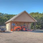 A small tan shed with an open front houses an orange tractor and various tools. A utility trailer is parked outside on a gravel lot, with green trees and blue sky in the background.