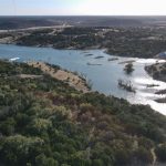 A river winds through a landscape of green trees and shrubs, with small islands and narrow peninsulas. A dirt road runs parallel to the shoreline, and scattered buildings are visible in the distance under a clear sky.