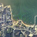 Aerial view of a lakeside residential area with houses, streets, docks extending into the water, and a marina with blue-roofed boat slips along the shoreline. Dense trees and curved roads surround the lake.