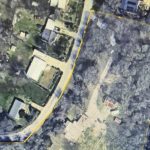 Aerial view of a residential area with houses, trees, and a curved road; a yellow outline marks the boundary of a large property containing multiple structures and wooded areas.