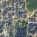 Satellite view of a suburban neighborhood with curved and straight streets, houses, trees, and docks along the edge of a body of water at the top right. Yellow dots highlight a section of the area.