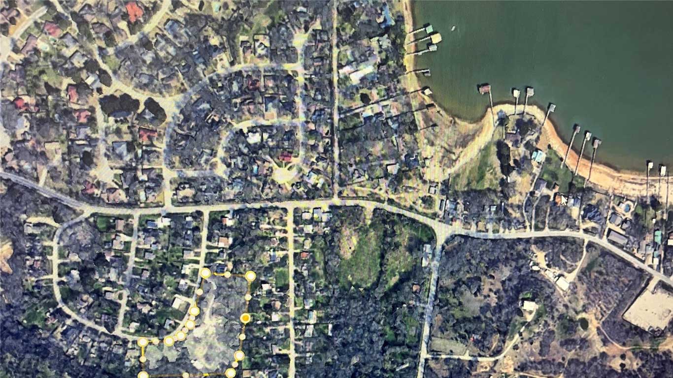 Satellite view of a suburban neighborhood with curved and straight streets, houses, trees, and docks along the edge of a body of water at the top right. Yellow dots highlight a section of the area.