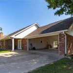 A brick house with an attached open two-car garage, concrete driveway, and a large yard with grass and trees under a clear blue sky.