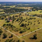 Aerial view of a large rural property outlined in white, featuring open grassy fields, scattered trees, and several buildings, surrounded by other fields and wooded areas.