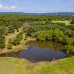 A small pond surrounded by green grass and dense trees in a rural landscape, with hills and a partly cloudy blue sky visible in the background.