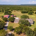 Aerial view of a rural property with a large house, two red barns, a driveway, and scattered trees surrounded by open green fields and distant hills under a blue sky.