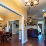 A warm, well-lit home interior features a hallway with a glass display cabinet, leading to a cozy living room with brown leather furniture and a fireplace, adjacent to a dining area with a wooden table and chandeliers.