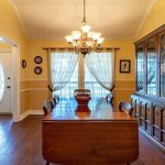 A dining room with yellow walls, a wooden table and chairs, a large china cabinet, chandelier, hardwood floors, and a window with sheer curtains. The entry door with glass panels is visible on the left.