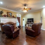 A cozy living room with brown leather chairs, a fireplace, wooden floors, a TV on a cabinet, and large windows with red plaid curtains. The room has warm lighting, decorative stars, and houseplants.