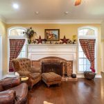 A cozy living room with a brick fireplace, brown leather chairs, wood flooring, plaid curtains, and decorative items on the mantle, including framed art and pottery. Warm lighting fills the room.