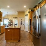 Spacious kitchen with wooden cabinets, granite countertops, stainless steel appliances, a large central island, tiled floor, and decorative greenery above cabinets. Natural light streams in from the left window.