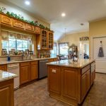 Spacious kitchen with wooden cabinets, granite countertops, central island, stainless steel appliances, tile flooring, and decorative greenery above the cabinets. Dining area visible through an open doorway.