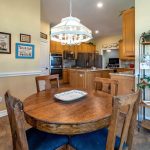A cozy kitchen with a round wooden dining table and four chairs, wood cabinetry, stainless steel appliances, wall art, a metal shelf with dishes and decor, and a hanging light fixture above the table.