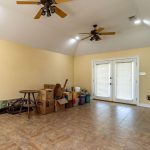 Spacious room with tiled floor, yellow walls, and two ceiling fans. Furniture includes a desk chair, side table, wooden cabinet, and shelves. Cardboard boxes and storage bins are against the wall near double doors with blinds.