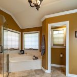 A bathroom with brown walls, a glass-enclosed shower, a bathtub with windows above, a towel hanging by the tub, a separate toilet area with a window, and a small chandelier on the ceiling.