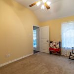 A small, cozy room with light yellow walls, beige carpet, a ceiling fan with lights, a window with sheer curtains, a rocking chair, and colorful quilts draped over chairs and a bench. An open door reveals an adjacent room.