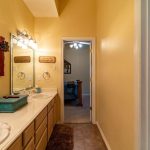 A bathroom with yellow walls, double sinks with a large mirror, decorative wall items, and a blue box on the counter. Through the open door, stairs and part of a living area are visible. A patterned shower curtain is on the right.