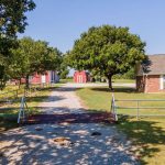 A gravel driveway with a metal gate leads to a red barn and a house with white trim surrounded by green trees and grass under a clear blue sky.