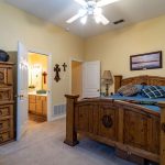 A cozy bedroom with rustic wooden furniture, including a bed, dresser, and desk. The bed is neatly made with blue and plaid bedding. Wall art, crosses, and a ceiling fan decorate the light yellow room.