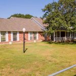 A single-story brick house with a large front porch, several windows, and a sloped roof, surrounded by grass, trees, and a metal fence under a clear blue sky.