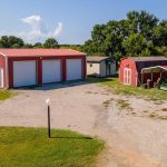 Aerial view of a rural property with a large red metal garage with three white doors, a smaller red shed with a covered area sheltering a green tractor, and grassy fields with scattered trees in the background.