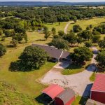 Aerial view of a rural property with a single-story house, large red barn, and small red shed surrounded by trees, open grassy fields, and a winding driveway under a clear sky.
