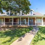A single-story brick house with a large front porch, white railings, and dormer windows. A curved sidewalk leads to the front door, and the yard features green grass and a large tree.