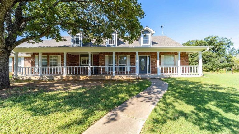 A single-story brick house with a large front porch, white railings, and dormer windows. A curved sidewalk leads to the front door, and the yard features green grass and a large tree.