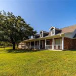 A red brick house with white trim and a wraparound porch sits on a large, grassy lawn under a clear blue sky. A mature tree provides shade nearby, and a white fence runs along the property.