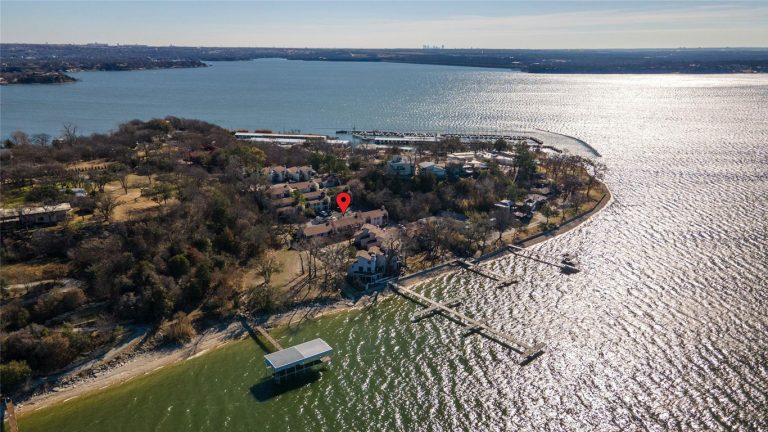 Aerial view of a peninsula with houses surrounded by water, a dock extending into the lake, and a red location marker above one house. The shoreline is lined with trees and the city skyline is visible in the distance.