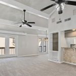 Spacious living area with vaulted ceiling, two ceiling fans, French doors to a balcony, carpeted floor, and an open kitchen with a breakfast bar and tile backsplash. Dining area with large window is visible.