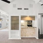 Modern open-concept living area with beige carpet, white walls, a built-in shelf, ceiling fans, and a small kitchen with stainless steel appliances and a tiled counter. Large windows provide ample natural light.