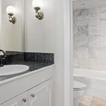 A bathroom with a white vanity, black granite countertop, and a sink with a black faucet. There’s a large mirror, wall-mounted light fixtures, and a bathtub with white and gray marble tile in the adjacent room.