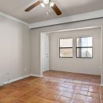 A bright, empty room with terracotta tile flooring, beige walls, white trim, two windows, a ceiling fan with lights, and a doorway leading to another area. Exposed wires are visible near one wall.