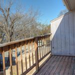 A small wooden balcony with a weathered floor and railing overlooks a residential area with leafless trees and neighboring houses under a clear blue sky.