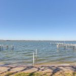 A calm lake under a clear blue sky with two wooden docks extending into the water from the shore. Shadows of tree branches are visible on the ground in the foreground.