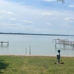 A peaceful lakeside scene with two wooden docks extending over calm water, grassy lawn in the foreground, and trees framing the view under a partly cloudy sky.