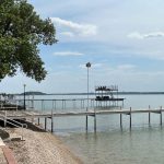 A lakeside scene with wooden docks extending over calm water, a tree providing shade on the left, patio chairs and tables along a paved walkway, and a two-level diving platform in the distance under a partly cloudy sky.