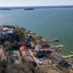 Aerial view of lakeside homes and docks along the shoreline, with a red location pin marking a specific building. The expansive lake dominates the right side of the image, with trees and houses on the left.