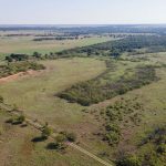 Aerial view of a vast rural landscape with grassy fields, scattered trees, dirt roads, and distant hills under a clear sky. A few small buildings are visible in the far distance.