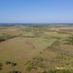 A wide aerial view of rolling, grassy fields with scattered trees under a clear blue sky, stretching to the horizon. No buildings are visible; the landscape appears rural and open.