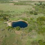 Aerial view of a small, circular pond surrounded by green grass, trees, and shrubs in a rural landscape, with open fields and a few scattered buildings in the distance.