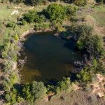 A small, dark pond surrounded by green trees and shrubs in a grassy, slightly hilly landscape, seen from above on a sunny day.
